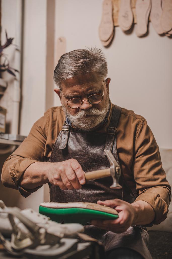 senior, bearded cobbler repairing shoe with hummer in workshop - Photo, Image