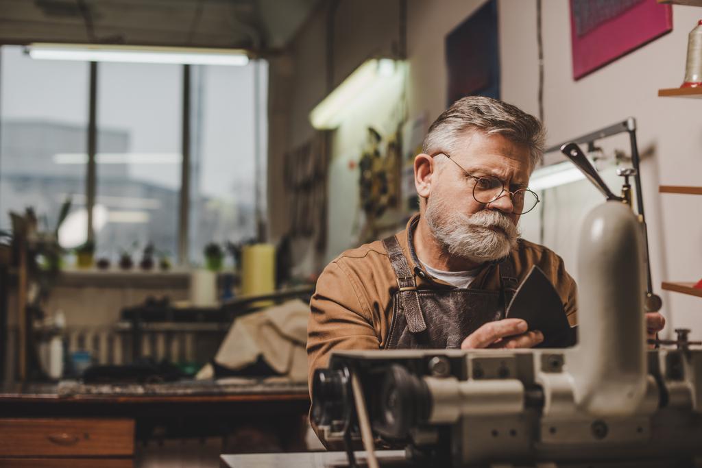 sénior, zapatero barbudo coser cuero en la máquina de coser en el taller
 - Foto, Imagen