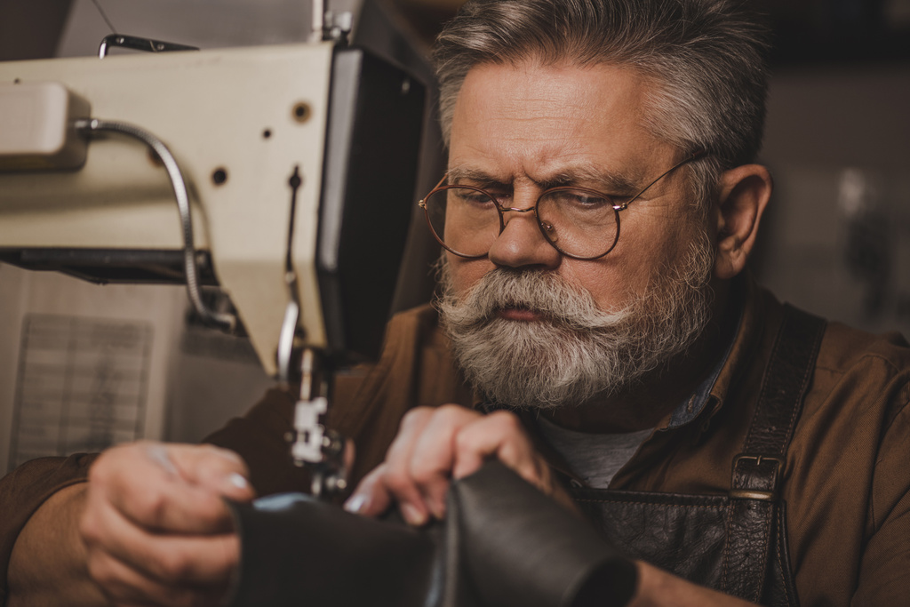 старший, бородатый сапожник шьет кожу на швейной машинке
 - Фото, изображение