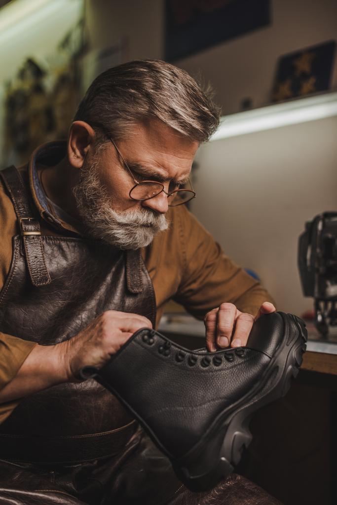 внимательный, старший сапожник держа кожаный сапог в мастерской
 - Фото, изображение