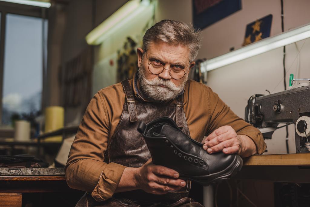 serious, senior shoemaker holding leather boot while looking at camera - Photo, Image