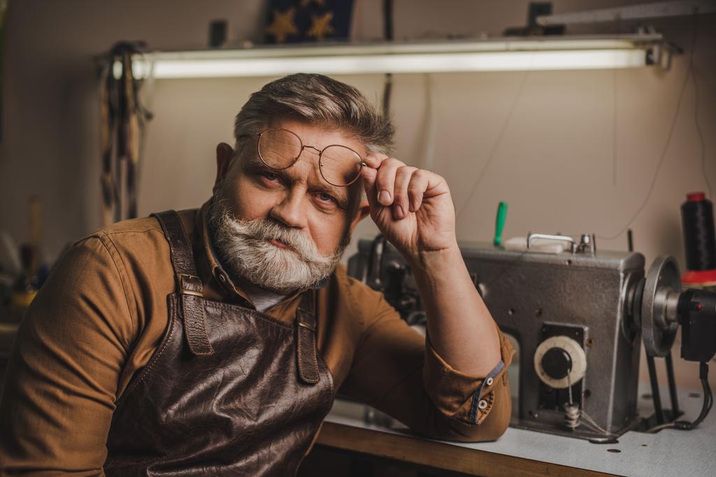 senior, confident cobbler looking at camera while sitting near sewing machine in workshop - Photo, Image