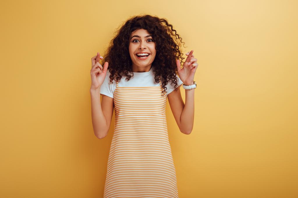 vzrušený bi-rasové dívka drží zkřížené prsty při úsměvu na fotoaparátu na žlutém pozadí - Fotografie, Obrázek