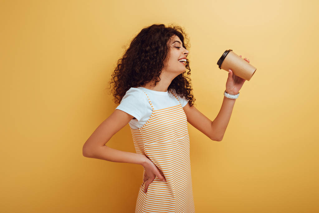 Радостная двурасовая девушка пьет кофе, стоя с рукой на бедре на желтом фоне
 - Фото, изображение
