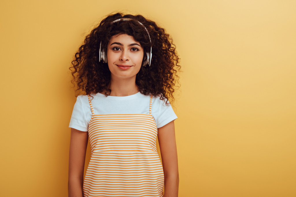όμορφη, χαμογελαστή bi-φυλετική κοπέλα σε ασύρματα ακουστικά κοιτάζοντας κάμερα σε κίτρινο φόντο - Φωτογραφία, εικόνα