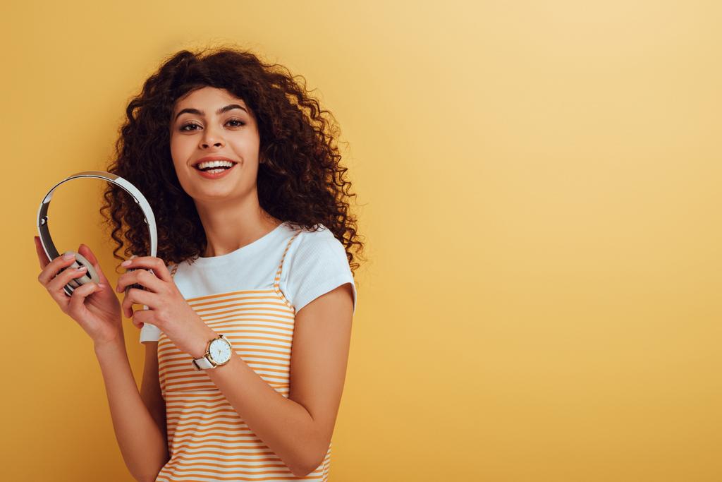 χαρούμενη bi-φυλετική κοπέλα χαμογελώντας στην κάμερα, ενώ κρατώντας ασύρματα ακουστικά σε κίτρινο φόντο - Φωτογραφία, εικόνα