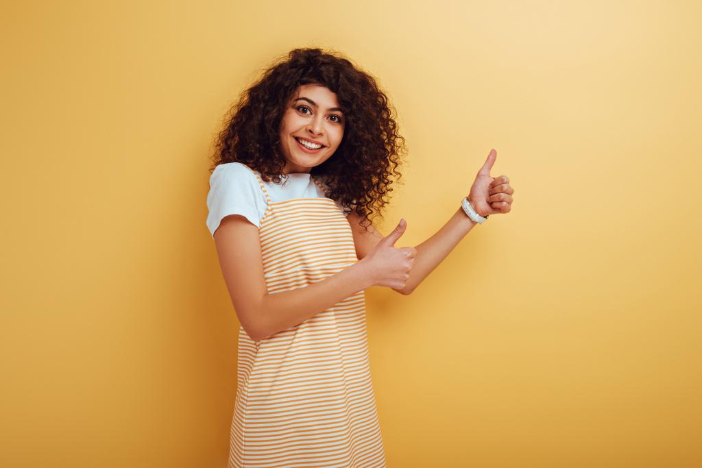 веселая девушка смешанной расы показывает большие пальцы вверх, глядя на камеру на желтом фоне
 - Фото, изображение