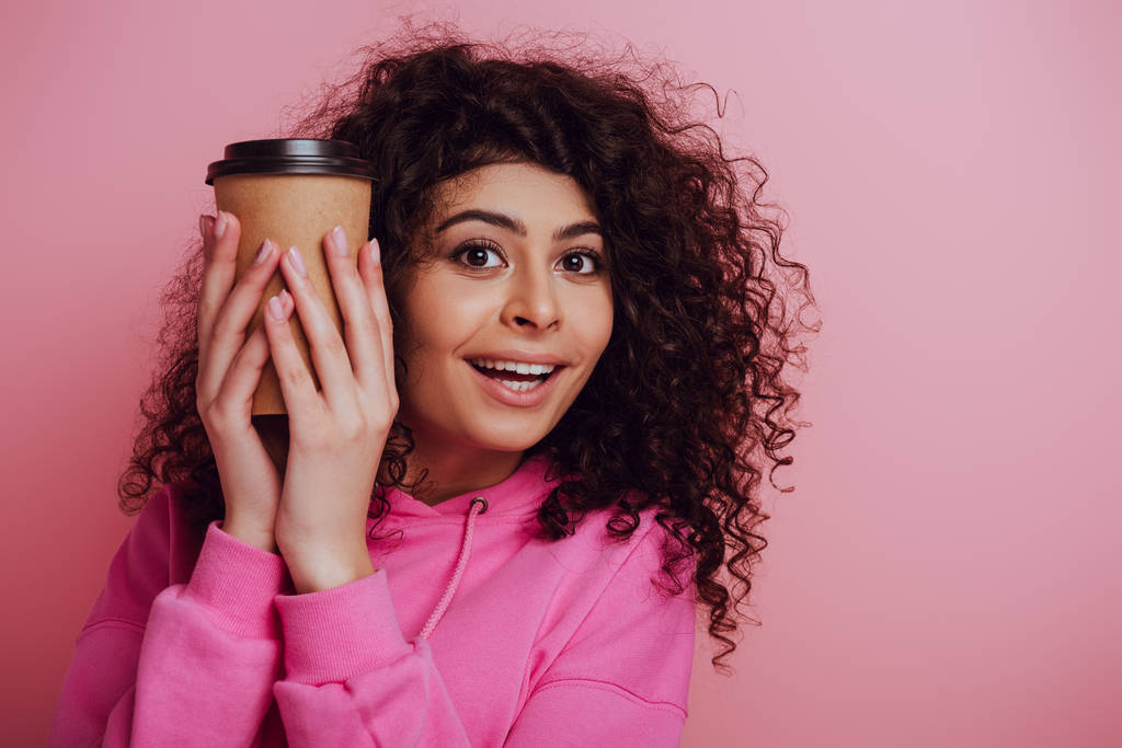 χαρούμενα bi-φυλετικό κορίτσι κρατώντας καφέ για να πάει, ενώ χαμογελά στην κάμερα σε ροζ φόντο - Φωτογραφία, εικόνα