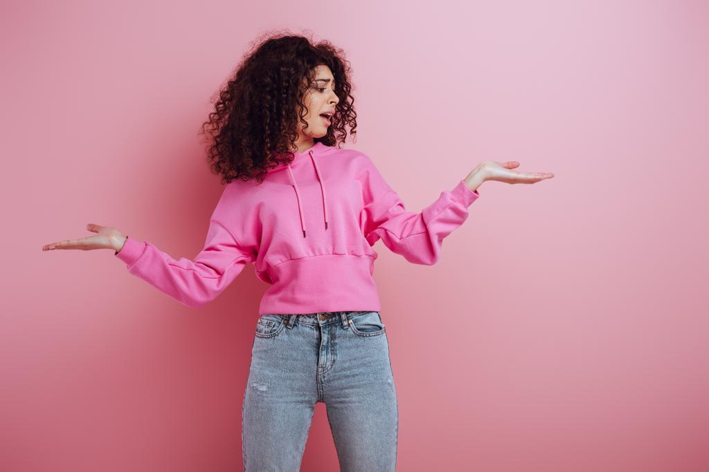displeased bi-racial girl standing with open arms on pink background - Photo, Image