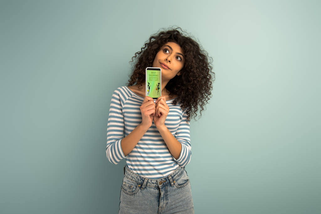 вдумчивая девушка смешанной расы смотрит в сторону, показывая смартфон с лучшим приложением для покупок на сером фоне
 - Фото, изображение