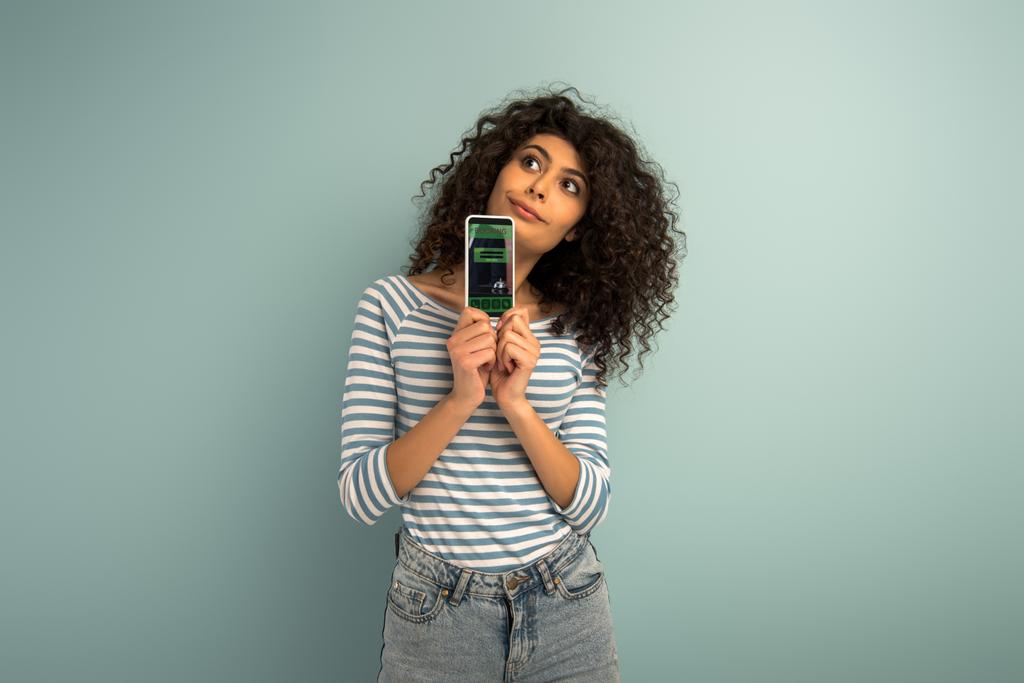 reflexiva chica de raza mixta mirando hacia otro lado mientras muestra el teléfono inteligente con la aplicación de reserva en fondo gris
 - Foto, Imagen