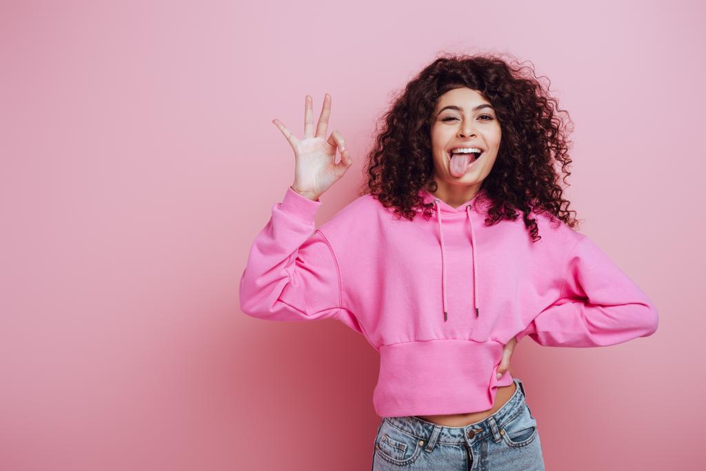 веселая двурасовая девушка, торчащая языком и показывающая нормальный жест на розовом фоне
 - Фото, изображение