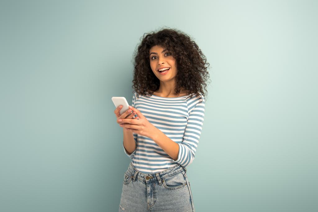 boldog bi-rasszista lány chat okostelefon, miközben nézi a kamera szürke háttér - Fotó, kép