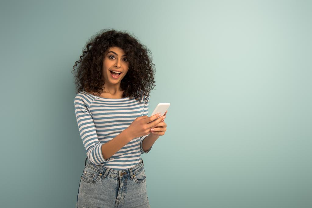 fröhliches zweirassiges Mädchen, das auf dem Smartphone chattet, während es in die Kamera auf grauem Hintergrund schaut - Foto, Bild
