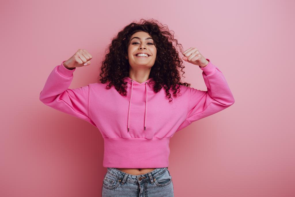 excited bi-racial girl showing winner gesture while smiling at camera on pink background - Photo, Image