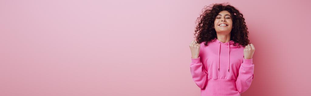 panoramic shot of excited bi-racial girl showing winner gesture while standing with closed eyes on pink background - Photo, Image