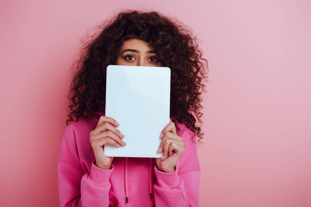 young bi-racial woman looking at camera while obscuring face with digital tablet on pink background - Photo, Image