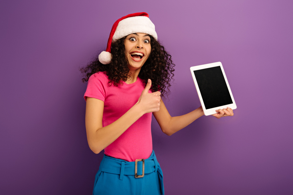 збуджена бі-расова дівчина в капелюсі Санта, що показує великий палець вгору, тримаючи цифровий планшет з порожнім екраном на фіолетовому фоні
 - Фото, зображення