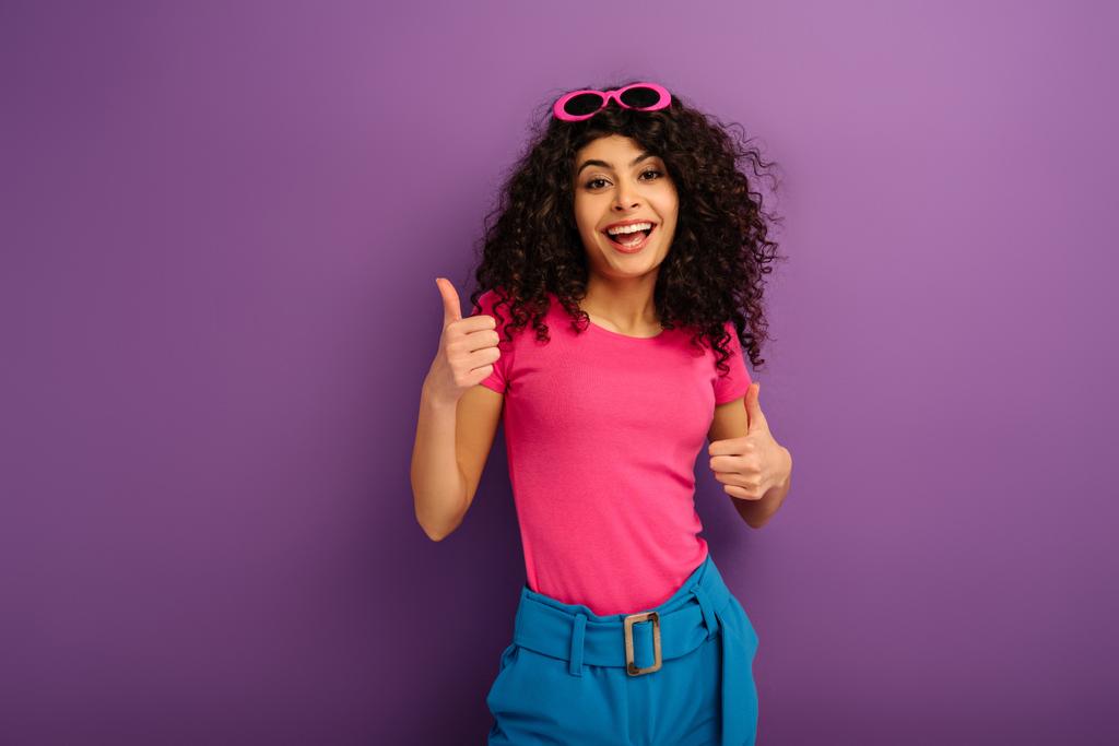 cheerful bi-racial girl showing thumbs up while smiling at camera on purple background - Photo, Image