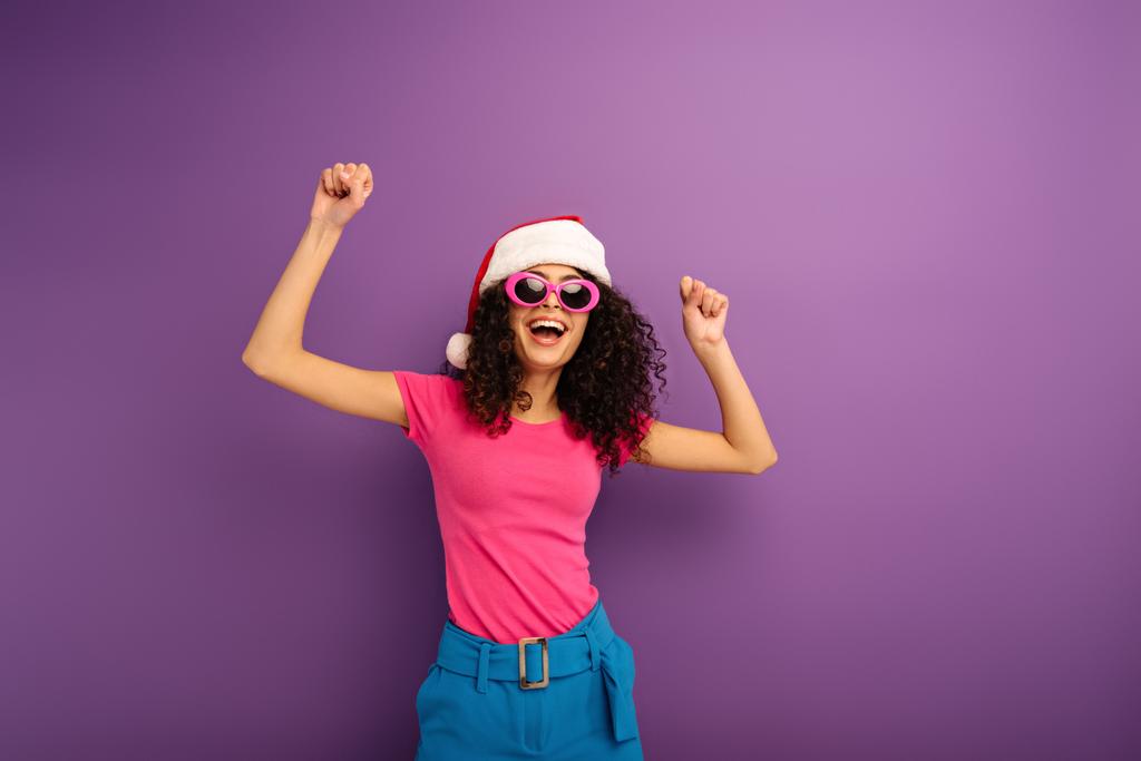 joyeuse fille bi-raciale en santa chapeau et lunettes de soleil dansant sur fond violet
 - Photo, image