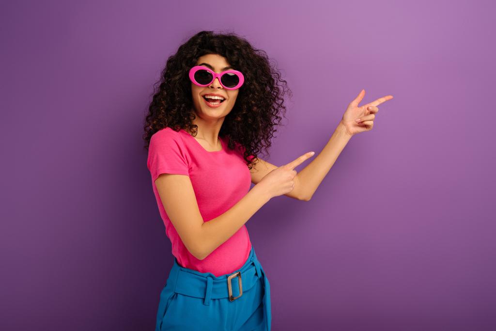 šťastný bi-rasové dívka v slunečních brýlích při pohledu na kameru, zatímco ukazuje prsty na fialové pozadí - Fotografie, Obrázek