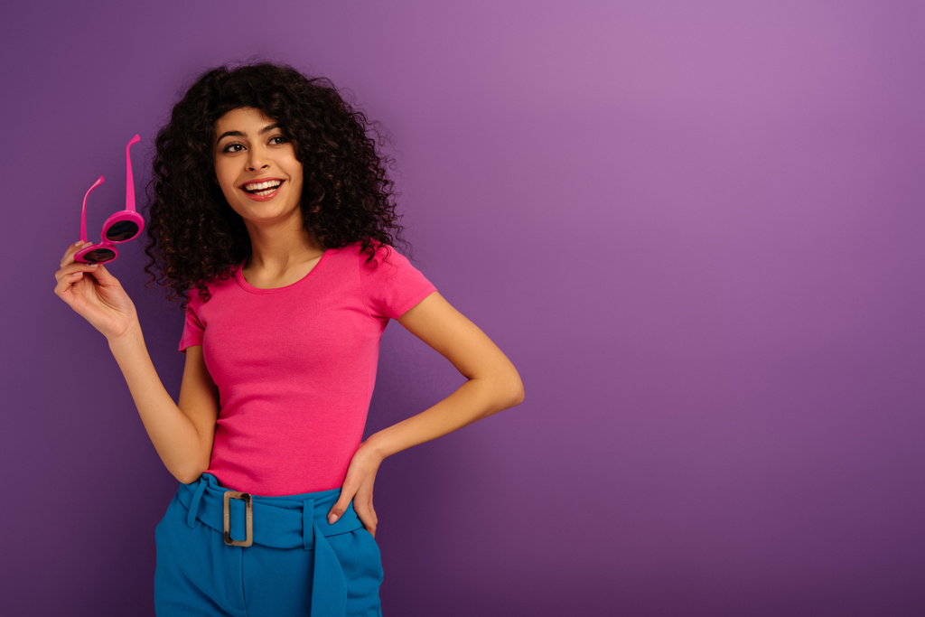 cheerful bi-racial girl holding sunglasses while looking away on purple background - Photo, Image