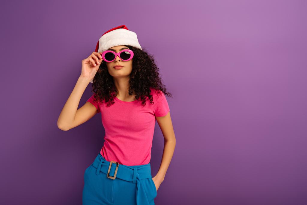 stylish mixed race girl in santa hat touching glasses while holding hand in pocket on purple background - Photo, Image