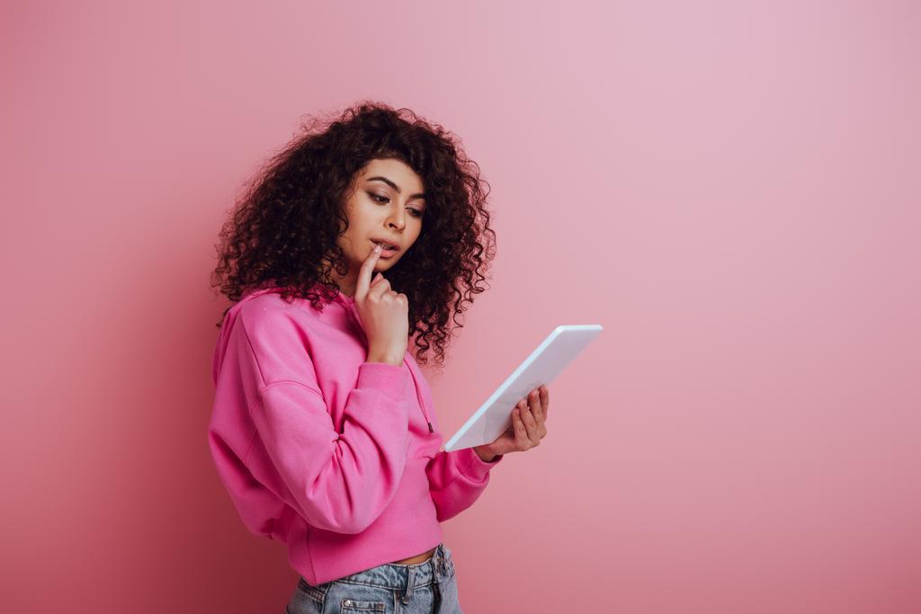 pensativo bi-racial chica tocando el labio mientras se utiliza la tableta digital sobre fondo rosa
 - Foto, imagen