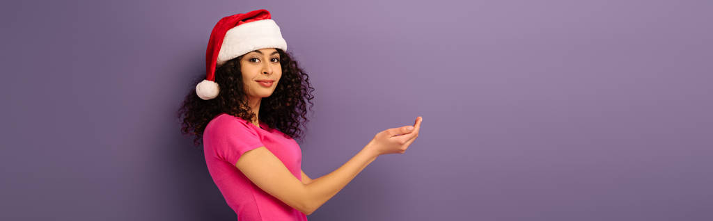 plano panorámico de chica de raza mixta sonriente en sombrero de santa de pie con las manos dobladas sobre fondo púrpura
 - Foto, imagen