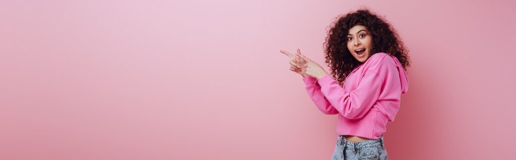 panoramisch schot van geschokt bi-raciale meisje kijken naar camera terwijl wijzend met vingers op roze achtergrond - Foto, afbeelding