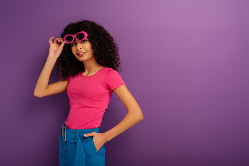 ładna bi-rasowy dziewczyna dotykając okulary podczas uśmiechania się do kamery na fioletowym tle - Zdjęcie, obraz
