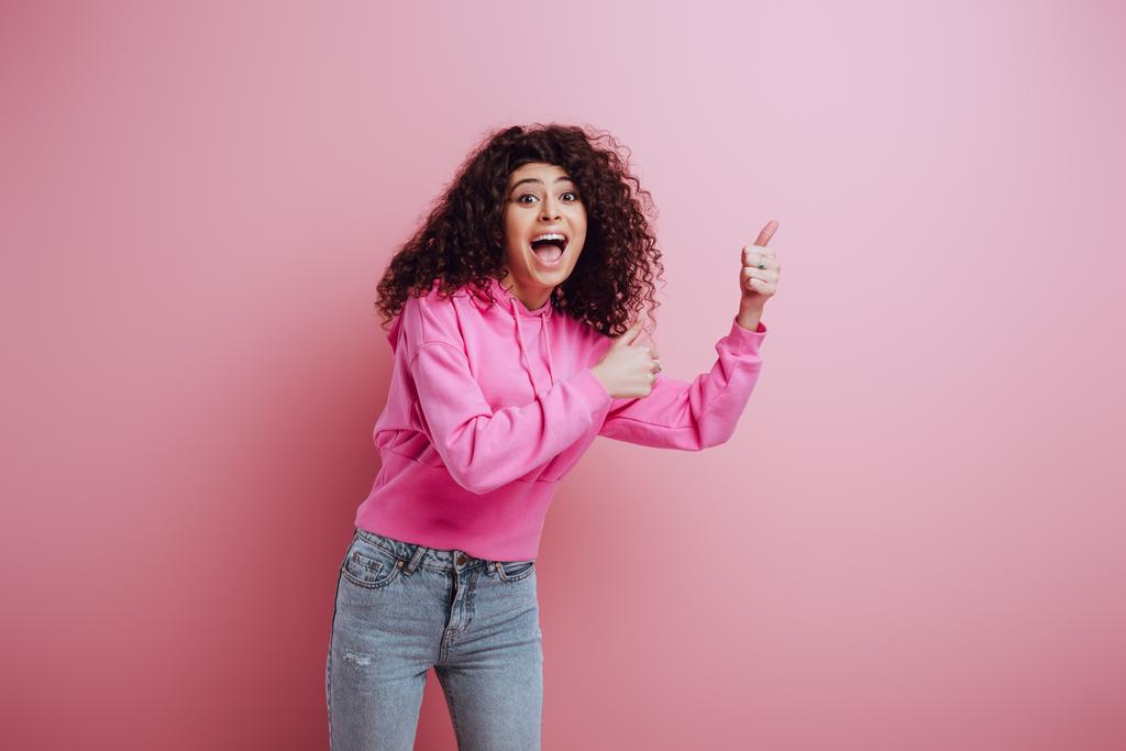  ενθουσιασμένοι bi-φυλετικό κορίτσι δείχνει αντίχειρες επάνω, ενώ κοιτάζοντας κάμερα σε ροζ φόντο - Φωτογραφία, εικόνα