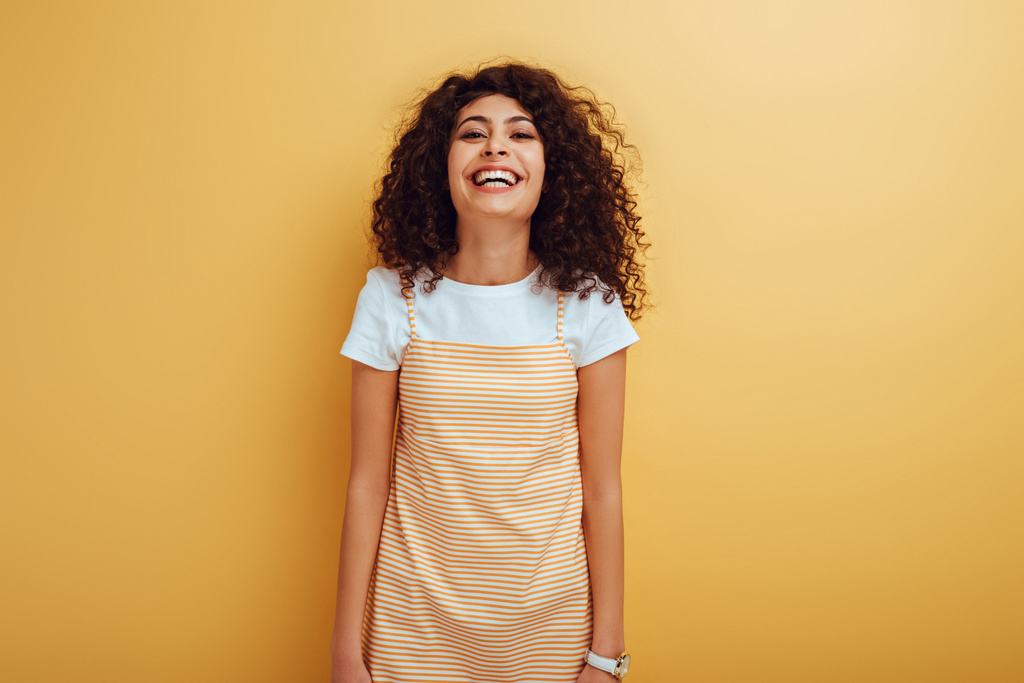 Смеющаяся девушка смешанной расы смотрит в камеру на желтом фоне
 - Фото, изображение