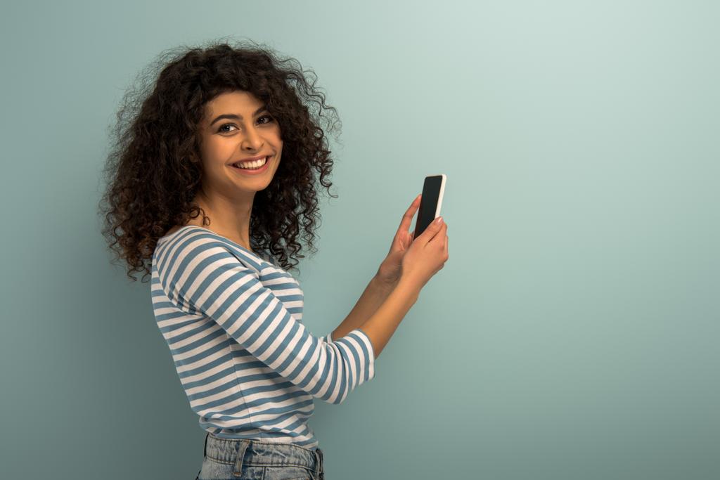 gelukkig bi-raciale meisje glimlachen op camera terwijl het houden van smartphone met leeg scherm op grijze achtergrond - Foto, afbeelding