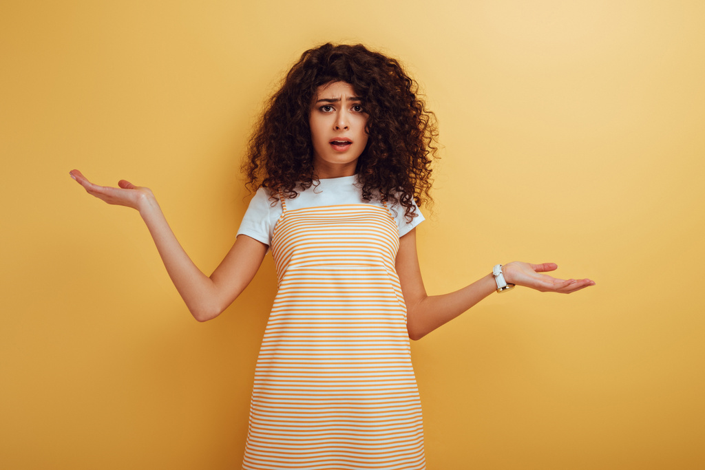 shocked, offended mixed race girl looking at camera while standing with open arms on yellow background - 写真・画像