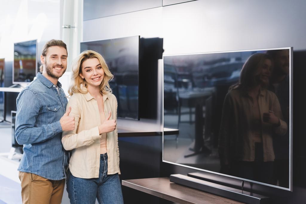 smiling boyfriend and girlfriend showing thumbs up in home appliance store  - Photo, Image