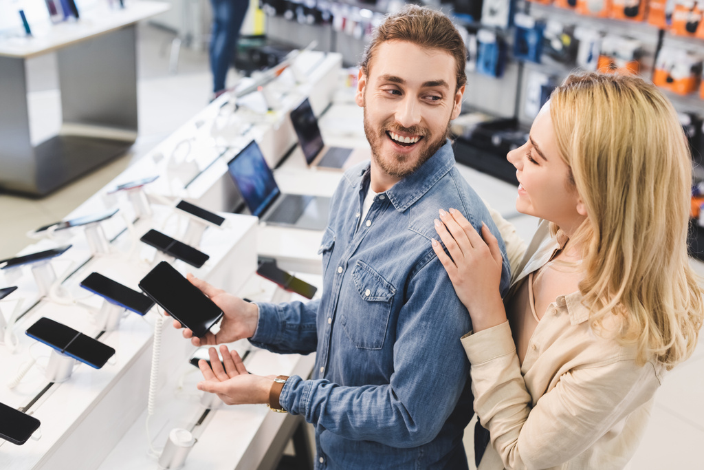 novio sonriente sosteniendo un nuevo teléfono inteligente y hablando con su novia en la tienda de electrodomésticos
  - Foto, Imagen