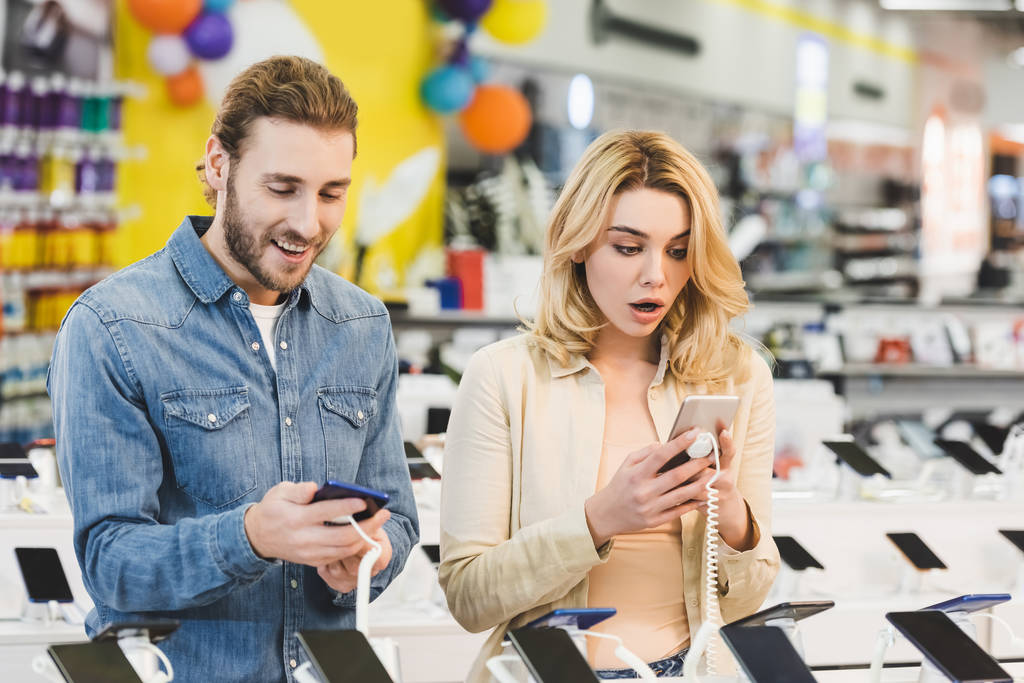 shocked boyfriend and girlfriend looking at new smartphones in home appliance store  - Photo, Image