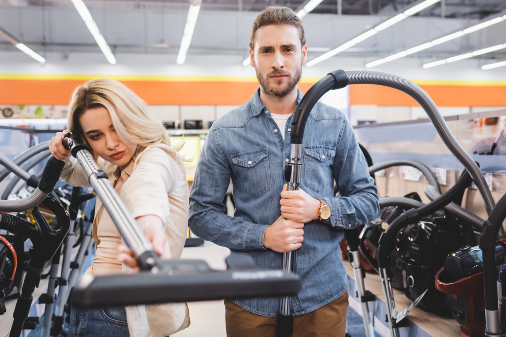 serious boyfriend and girlfriend holding vacuum cleaners in home appliance store  - Photo, Image