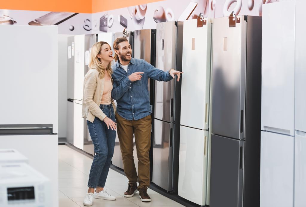 smiling girlfriend and boyfriend pointing with fingers at fridge in home appliance store  - Photo, Image
