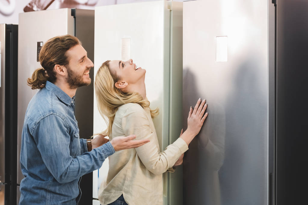 vista lateral do namorado sorridente apontando com a mão e namorada tocando geladeira na loja de eletrodomésticos
  - Foto, Imagem