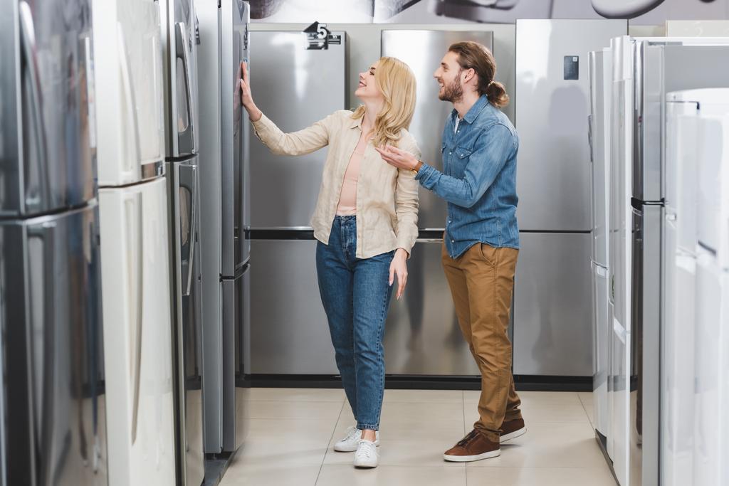 χαμογελαστός φίλος δείχνει με το χέρι και τη φίλη αγγίζοντας ψυγείο στο κατάστημα οικιακών συσκευών  - Φωτογραφία, εικόνα