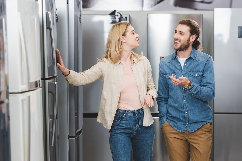 χαμογελαστός φίλος δείχνει με το χέρι και τη φίλη αγγίζοντας ψυγείο στο κατάστημα οικιακών συσκευών  - Φωτογραφία, εικόνα