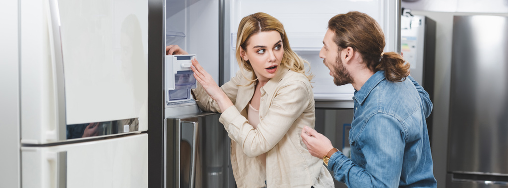 Panoramaaufnahme von schockiertem Freund und Freundin, die neben Kühlschrank in Haushaltsgerätegeschäft stehen  - Foto, Bild