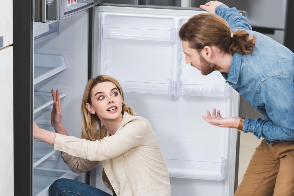 Freund spricht mit geschockter Freundin in der Nähe von Kühlschrank in Haushaltsgerätegeschäft  - Foto, Bild