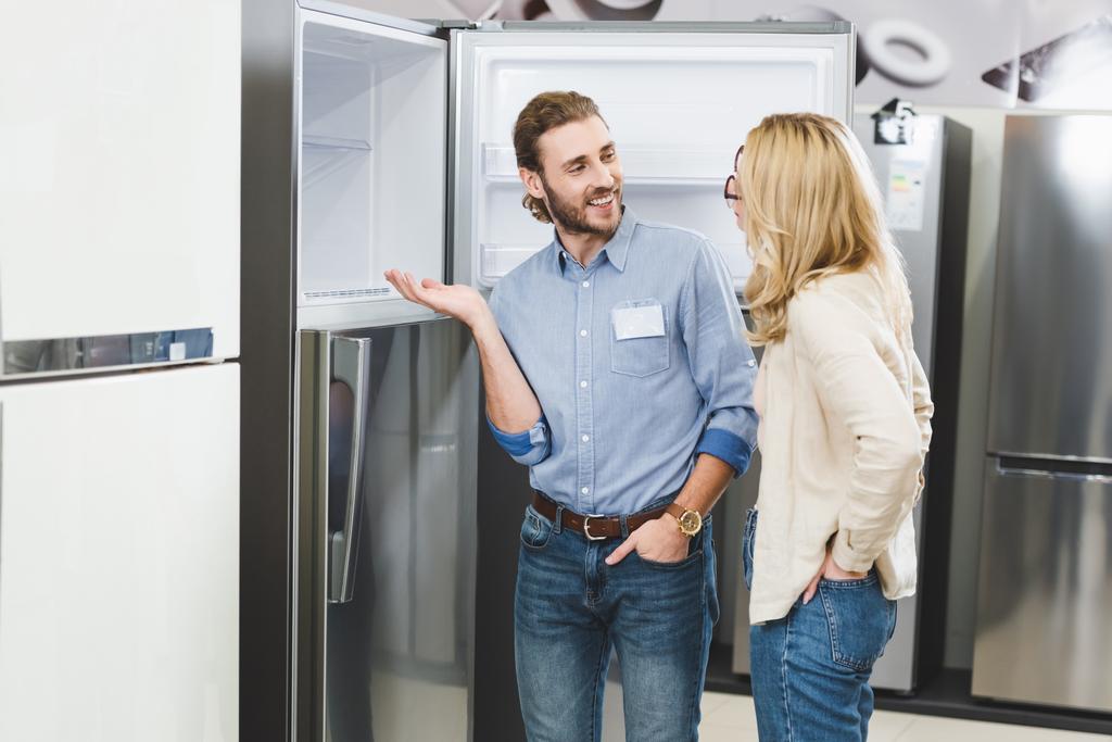 usmívající se konzultant ukazuje rukou na ledničku a mluví se ženou v obchodě s domácími spotřebiči  - Fotografie, Obrázek