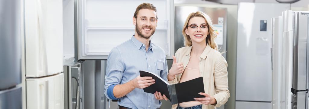 панорамний знімок усміхненого консультанта, який тримає папку і жінку, що показує, як в магазині побутової техніки
  - Фото, зображення