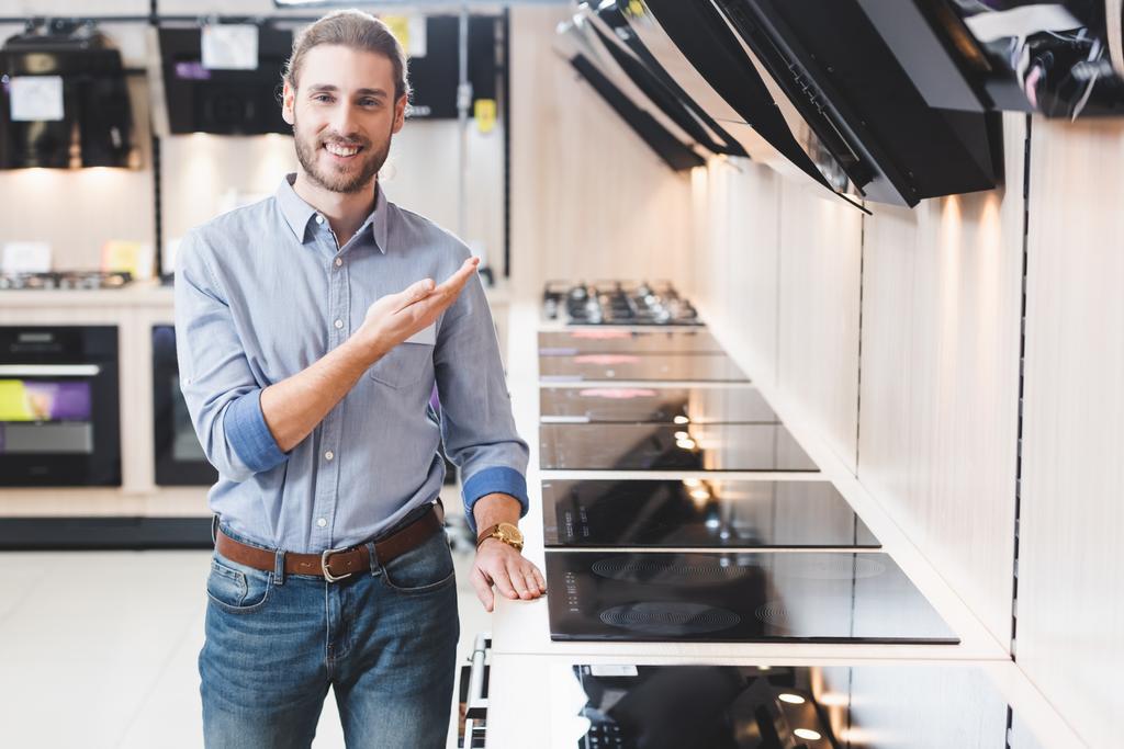 uśmiechnięty konsultant wskazujący ręką na okapy kuchenki w sklepie AGD - Zdjęcie, obraz