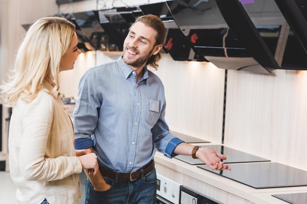 uśmiechnięty konsultant rozmawiający z kobietą w pobliżu okapów kuchennych i kuchenek w sklepie AGD  - Zdjęcie, obraz
