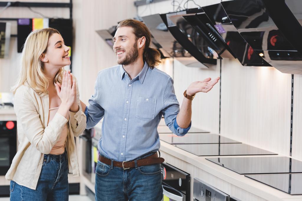 χαμογελαστός σύμβουλος δείχνει shrug χειρονομία και η γυναίκα δείχνει παρακαλώ χειρονομία κοντά απορροφητήρες κουζίνα στο κατάστημα οικιακών συσκευών  - Φωτογραφία, εικόνα